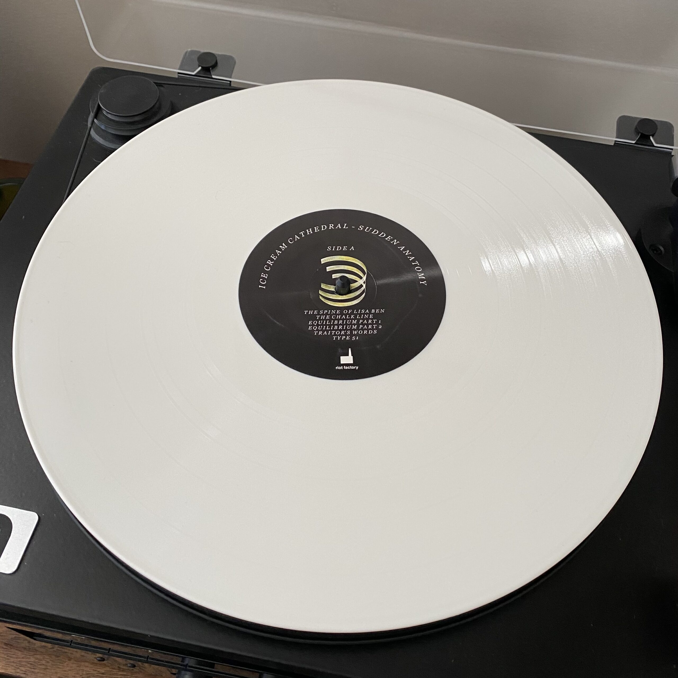 Image description: Ice Cream Cathedral's "Sudden Anatomy" vinyl album is on a record player. The album is white and has a black circle at the middle that shows the titles for Side A. "The Spine of Lisa Ben" is the first track on Side A.