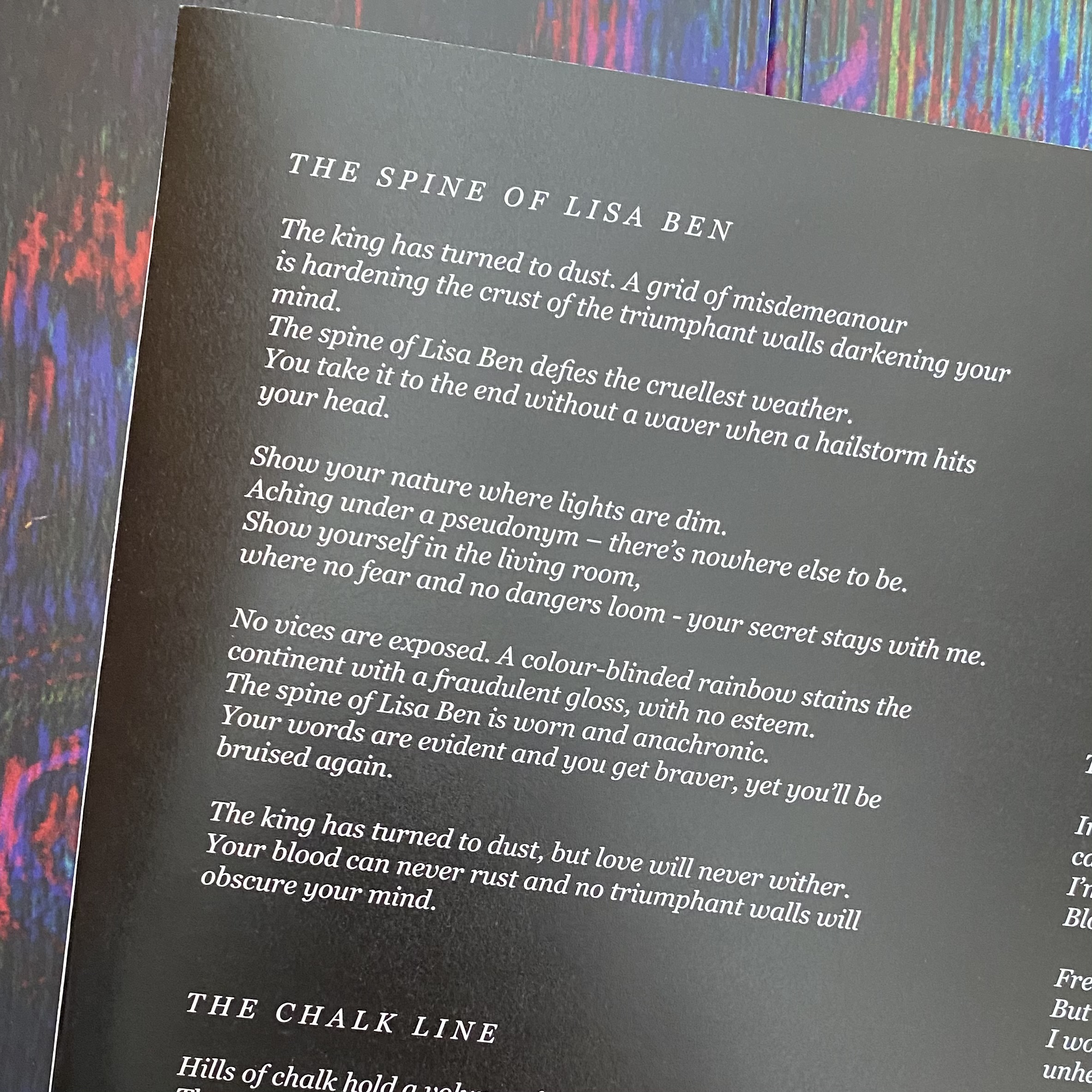 Image description: A photograph of the interior paper sleeve from the vinyl album "Sudden Anatomy," with the multicolored cardboard cover for the album in the background. The paper is black and the lyrics are printed in white italicized serif font. The lyrics are as follows:  The king has turned to dust. A grid of misdemeanour is hardening the crust of the triumphant walls darkening your mind. The spine of Lisa Ben defies the cruellest weather. You take it to the end without a waver when a hailstorm hits your head.  Show your nature where lights are dim. Aching under a psudonym — there's nowhere else to be. Show yourself in the living room, where no fear and no dangers loom — your secret stays with me.  No vices are exposed. A colour-blinded rainbow stains the  continent with a fraudulent gloss, with no esteem. The spine of Lisa Ben is worn and anachronic. Your words are evident and you get braver, yet you'll be bruised again.  The king has turned to dust, but love with never wither.  Your blood can never rust and no triumphant walls will obscure your mind.